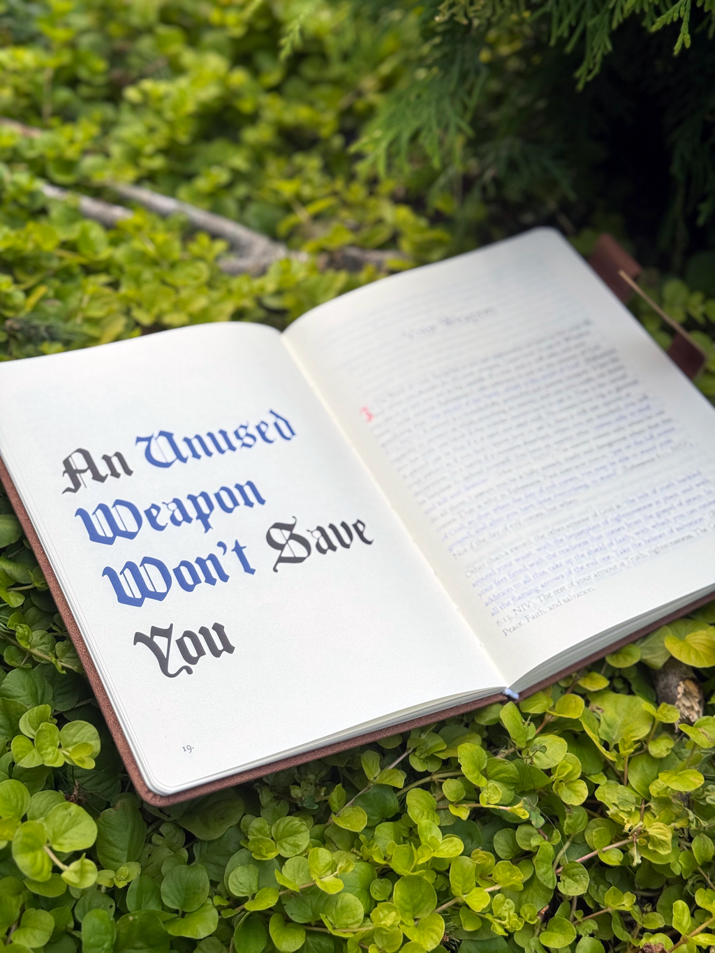 The Jesus Morning Journal opened lying on green foliage, with the title page reading 'An Unused Weapon Won't Save You' in black and blue gothic-style lettering.