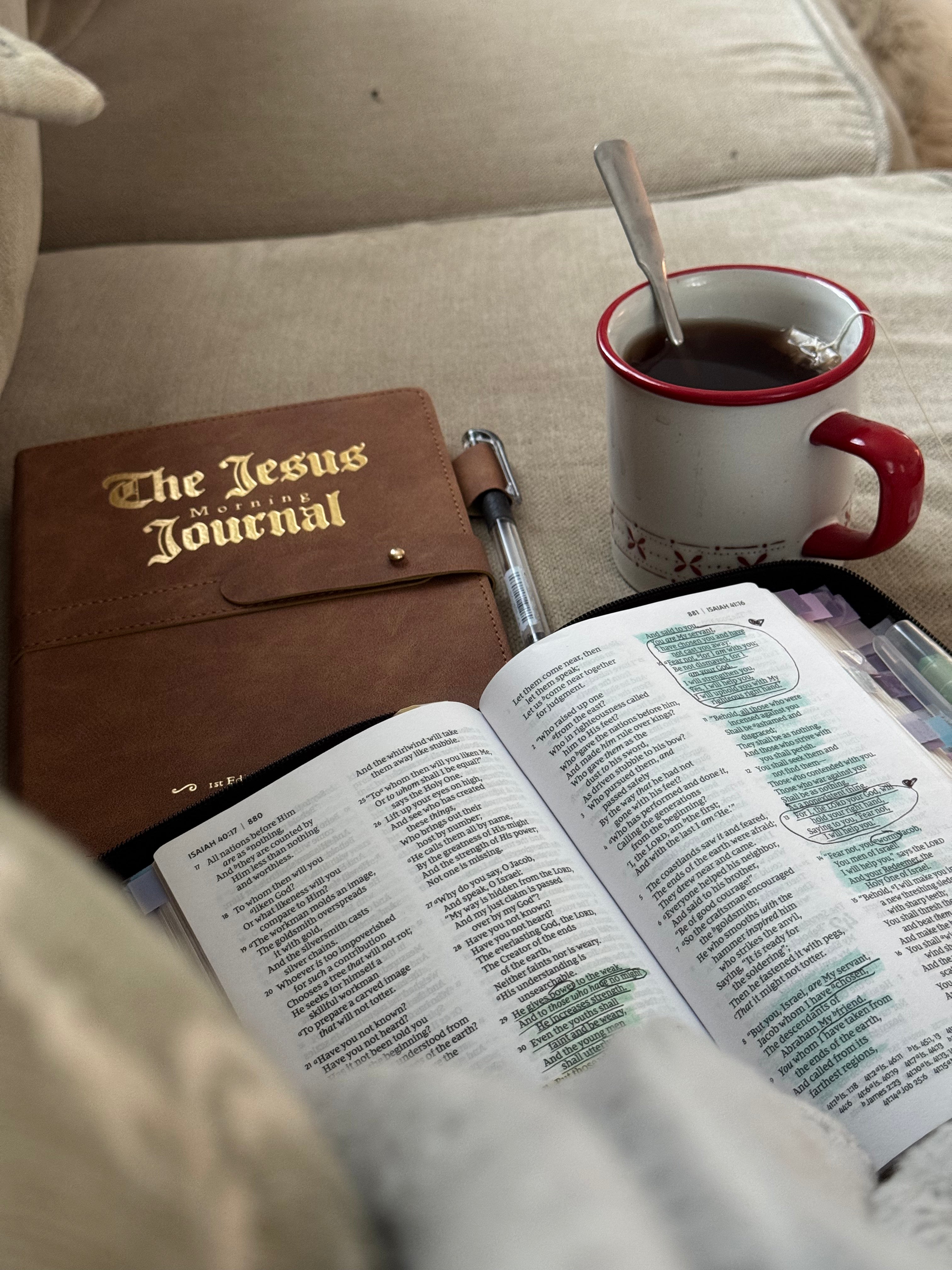 A brown leather journal with gold-embossed text reading 'The Jesus Journal' lies next to an open Bible and a white mug with a red rim on a beige couch.